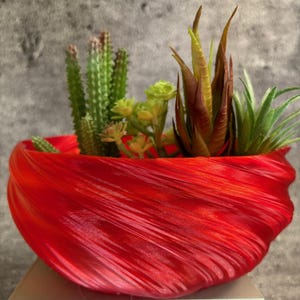May include: A vibrant red, decorative planter filled with a variety of artificial succulents and cacti. The planter has a unique, swirling design. The arrangement includes green cacti, yellow-green succulents, and brown and green plants.