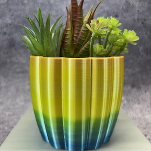 May include: A decorative planter with a gradient design, transitioning from yellow to blue. The fluted pot holds a variety of artificial succulents in shades of green and brown. The planter is approximately 5 inches tall.