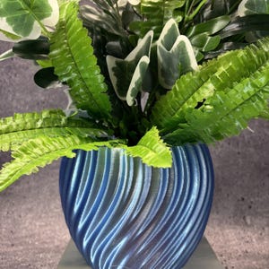 May include: A metallic blue planter with a spiralling, ribbed texture containing a lush arrangement of artificial green and white variegated ivy leaves and fern fronds. The plant pot features a distinct 3D-printed finish with a glossy sheen.