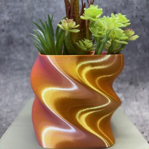 Puede incluir: Una maceta decorativa de forma abstracta, con un degradado de naranja, dorado y blanco. Contiene una variedad de suculentas y vegetación artificial. El diseño presenta una forma ondulada y retorcida, añadiendo un toque moderno a la decoración.