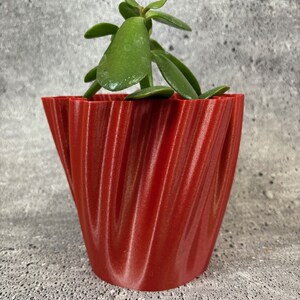 Può includere: Un vaso da fiori rosso ondulato stampato in 3D con una piccola pianta grassa verde al suo interno.