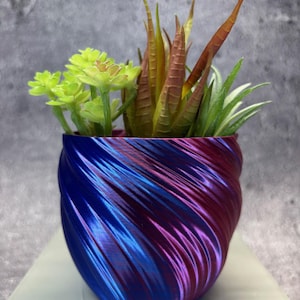 Puede incluir: Una pequeña maceta decorativa con un diseño arremolinado azul y morado, llena de una variedad de suculentas artificiales. Las suculentas incluyen plantas verdes, amarillas y de color marrón rojizo. La maceta está sobre una superficie blanca.