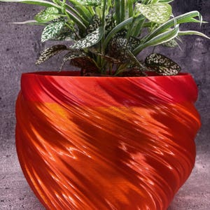 Pode incluir: Um vaso de plantas bicolor laranja e vermelho, impresso em 3D, com um design em espiral. O vaso contém uma planta verde exuberante com folhas salpicadas de branco e verde escuro. O vaso está sobre uma superfície cinza texturizada.