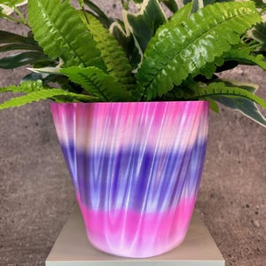 May include: A purple, pink and white swirl patterned planter with a green leafy plant inside. The planter is sitting on a light grey square pedestal.