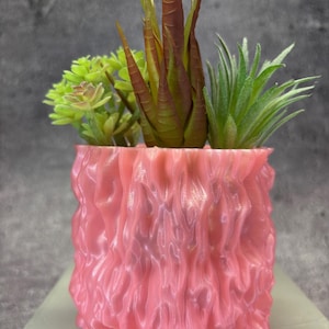 Puede incluir: Una maceta de cerámica rosa texturizada con tres suculentas artificiales. La maceta tiene un diseño ondulado y orgánico.