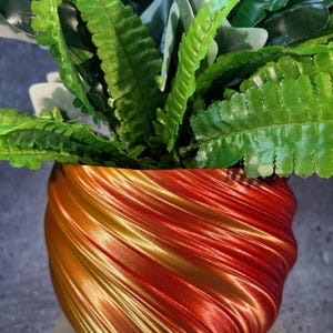 Op de afbeelding: Een rode en gouden swirl-pot met een groene, bladerrijke plant erin. De pot is gemaakt van een glad, glanzend materiaal.