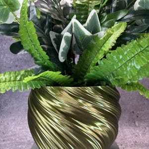 May include: A metallic olive green planter with a spiraling, ribbed texture containing a dense arrangement of artificial greenery. The plant features a mix of dark green leaves and lighter, variegated foliage with white edges, creating a full look.