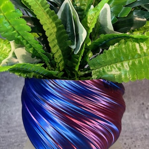 Może przedstawiać: Iryzujący niebieski i fioletowy doniczka z wirującym wzorem. Doniczka jest wypełniona zielonymi, liściastymi roślinami.