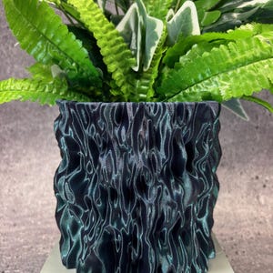 Op de afbeelding: Een donkergroene, getextureerde keramische pot met een groene, bladerrijke plant erin. De pot heeft een golvend, organisch ontwerp.