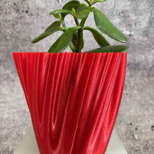 Puede incluir: Una maceta roja impresa en 3D con un diseño ondulado y texturado. Una pequeña planta verde está creciendo dentro de la maceta.