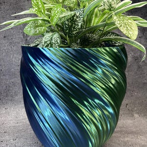 May include: A plant pot with a metallic, iridescent finish that shifts between deep blue and vibrant green. The exterior features a distinct, diagonal spiral texture. A leafy green and white variegated plant is potted inside the container.