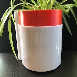 May include: A white and red plastic planter with a green plant growing inside. The planter has a unique design with a white outer layer and a red inner layer.