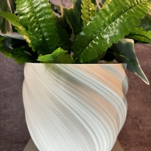 May include: A white, swirled-design planter pot filled with green and white artificial plants. The pot has a unique, textured pattern and sits on a light gray surface. The plants have vibrant green leaves.