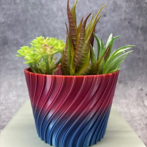 Puede incluir: Una maceta decorativa con un diseño en espiral, que transiciona del rojo al azul. La maceta contiene una variedad de suculentas artificiales en tonos verdes y marrones. La maceta está sobre una superficie blanca.