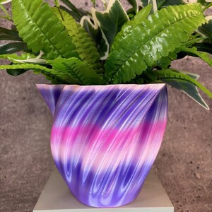 May include: A purple and pink swirl planter with a green leafy plant inside. The planter is made of a smooth, glossy material and has a unique, twisted design.