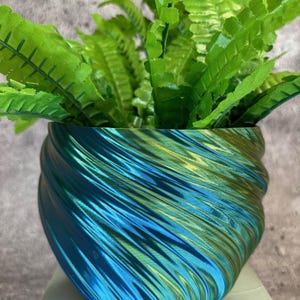May include: A vibrant, decorative planter with a swirling, iridescent blue and green finish. The planter holds a lush, artificial fern with bright green leaves. The pot sits on a white surface, creating a striking contrast.