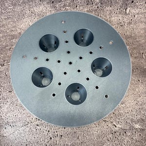 Op de afbeelding: Een cirkelvormig, grijs metalen object met vijf grote, kegelvormige openingen en talrijke kleinere gaten. Het object, met een matte afwerking, lijkt een onderdeel te zijn van een groter mechanisme, mogelijk voor industrieel gebruik. Diameter ongeveer 20 cm.
