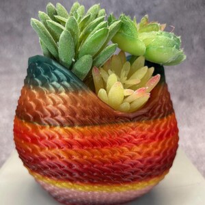 May include: A colourful, 3D printed planter with a textured, woven design in shades of red, orange, yellow, and green. The planter is filled with artificial succulents.