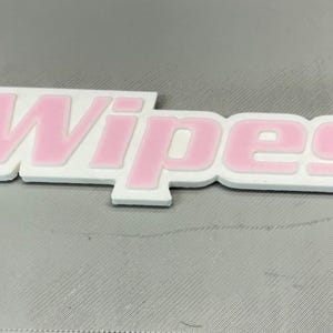 Puede incluir: Un letrero blanco y rosa que dice "Wipes" en una fuente negrita y redondeada. La palabra es rosa con un contorno blanco, sobre un fondo gris. El letrero parece ser una etiqueta de producto o un elemento de embalaje.