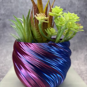 May include: A decorative planter with a swirling design in shades of blue and magenta, holding a variety of artificial succulents. The faux plants include green, yellow, and brown foliage, creating a vibrant contrast against the pot's colors.