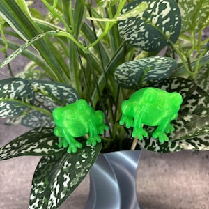 May include: A decorative arrangement featuring two bright green frog figurines nestled among artificial greenery. The leaves have a speckled pattern, and the vase is a light blue-gray with a twisted design. The frogs are approximately 1 inch tall.