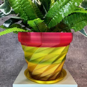 May include: A colorful, geometric-patterned flower pot with a matching saucer. The pot transitions from red at the top to yellow and brown at the bottom. The pot holds green leafy plants. The pot sits on a white surface.