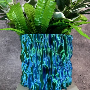 Op de afbeelding: Een blauwgroene, getextureerde plantenbak, gevuld met groen blad. De plantenbak heeft een opvallend, geribbeld ontwerp, waardoor het een opvallend decoratief stuk is. De bloempot staat op een wit oppervlak.
