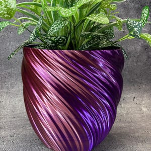 May include: A decorative plant pot with a swirling design in shades of purple and pink. The pot is filled with a green plant with white speckled leaves. The pot has a glossy finish and sits on a gray surface.