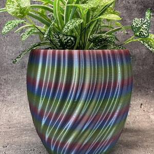 Op de afbeelding: Een decoratieve plantenpot met een wervelend patroon in groen, blauw en rood. De pot bevat een selectie van groene planten met wit en groen gespikkelde bladeren. De pot staat op een grijze ondergrond. De pot is ongeveer 20 cm hoog.