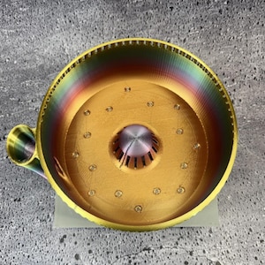 May include: A circular, 3D-printed coffee dripper featuring a gold-colored base with a central, raised, multi-slotted cone. The outer wall displays a vibrant, rainbow-colored gradient finish, and a small, matching handle extends from the side.