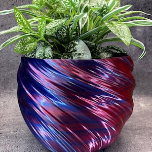 Op de afbeelding: Een levendige, gewervelde plantenbak in tinten rood, blauw en paars herbergt een weelderige groene plant met wit gespikkelde bladeren. Het ontwerp van de plantenbak heeft een getextureerde, glanzende afwerking, waardoor een dynamisch visueel effect ontstaat. De plant voegt een natuurlijk element toe aan de decoratieve pot.