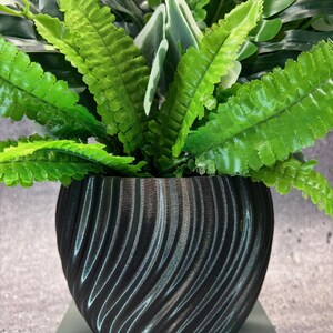 May include: A black, textured plant pot with a swirling design, filled with artificial green ferns and other foliage. The pot is round and sits on a light surface, contrasting with the dark pot. The pot's diameter is approximately 20 cm.