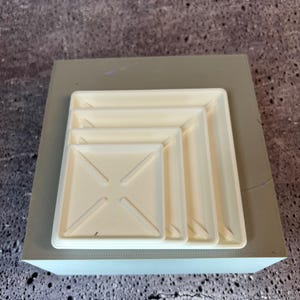 May include: Four white plastic square plant pots stacked on top of each other. The pots are all the same shape but different sizes.