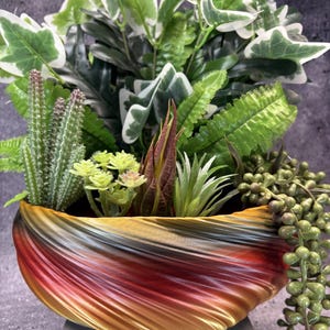 May include: A decorative bowl with a swirling, gradient finish transitioning from deep red to orange and gold. The bowl is filled with an assortment of artificial plants, including green ivy, a tall cactus, small succulents, and a trailing string of pearls.
