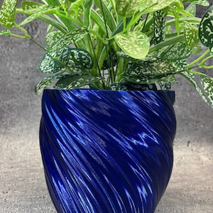 Puede incluir: Una maceta azul vibrante con un diseño en espiral texturizado, que contiene una exuberante planta verde con hojas moteadas de blanco. La planta tiene hojas largas y delgadas y una apariencia completa y saludable. El acabado brillante de la maceta añade un toque moderno.
