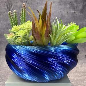Op de afbeelding: Een diepblauwe plantenpot met een metallic afwerking en een spiraalvormige, geribbelde textuur, gevuld met een assortiment kunstmatige vetplanten. De planten bestaan uit hoge groene cactussen, een centrale roodbruine vetplant en lichtgroen blad.