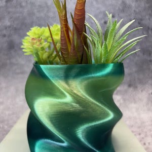 Pode incluir: Um vaso decorativo verde, com redemoinhos, cheio de suculentas artificiais. O vaso tem um brilho metálico e um design ondulado único. As suculentas incluem plantas verdes e avermelhadas.