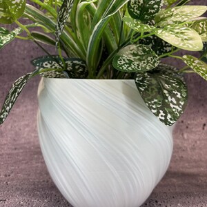 Op de afbeelding: Een witte, gewervelde keramische plantenpot met een levendige kamerplant. De plant heeft lange, groen-witte gestreepte bladeren en bladeren met witte vlekken. De pot heeft een afgeronde vorm met een textuur, gewerveld ontwerp.