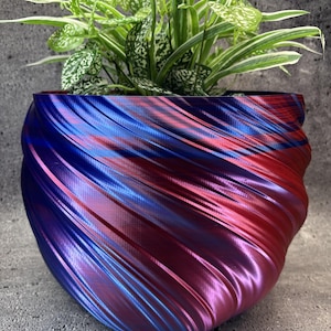Op de afbeelding: Een ronde, spiraalvormige plantenbak met een tweekleurige metallic afwerking die verschuift tussen levendig blauw en diep magenta. Het oppervlak heeft een textuur met ribbelpatroon. Een groen-witte bonte plant staat in de pot.