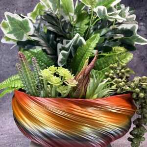 May include: A bowl-shaped planter with a swirling, multi-colored gradient finish in shades of orange, yellow, and dark brown. The planter is filled with an assortment of artificial greenery, including variegated ivy, ferns, small cacti, and succulents.