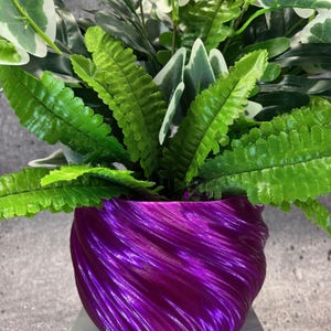 May include: A vibrant purple, textured planter filled with an assortment of green and white artificial plants. The planter has a swirling design and is filled with various types of faux foliage.