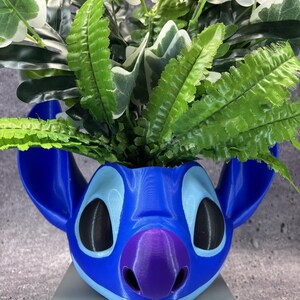 May include: A blue 3D-printed planter shaped like the head of the character Stitch, featuring large ears, a purple nose, and black eyes. The planter is filled with artificial green fern-like foliage and variegated leaves with white edges.