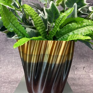 Può includere: Un vaso decorativo per piante con un design a spirale, con una sfumatura di colori dall'oro al marrone. Il vaso contiene una varietà di piante verdi con diverse forme di foglie, tra cui felci e edera. Il vaso è su una superficie bianca.