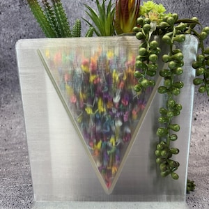 May include: A rectangular, silver-colored planter with a triangular cutout filled with colorful, abstract shapes. The planter holds various artificial succulents, including cacti and trailing green plants, creating a vibrant display.