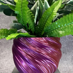 May include: A decorative plant pot with a swirling design in shades of purple and red. The pot holds a variety of green plants with different leaf shapes and textures. The pot is sitting on a light gray surface.