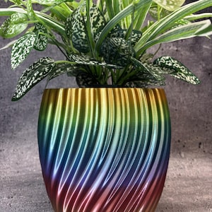 May include: A plant pot with a vertical spiral ribbed texture featuring a gradient of metallic colours including gold, blue, purple, and red. The pot contains a green and white variegated leafy plant with pointed foliage and speckled patterns.