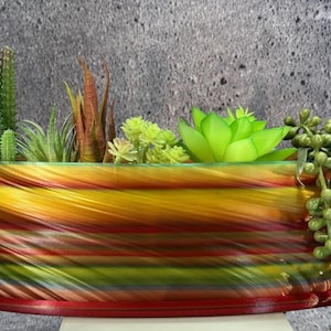 Op de afbeelding: Een ovale bloempot met een levendig, wervelend patroon in geel, oranje, rood en groen. Het bevat een selectie van kunstmatige vetplanten en cactussen, plus een hangplant met kleine groene kralen. Een decoratief item.