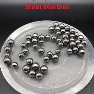 Pode incluir: Uma coleção de bolas de gude de aço brilhantes de vários tamanhos está espalhada em uma superfície circular cinza claro. O texto "Extra/Replacement Steel Marbles" é exibido em vermelho na parte superior. As bolas de gude têm uma aparência metálica reflexiva.