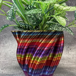 May include: A colorful, rainbow-striped, 3D printed planter with a green plant inside. The planter has a swirling, textured design and is made of plastic.