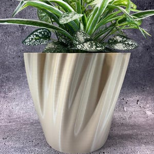 May include: A beige, spiral-textured planter pot holding a houseplant with green and white leaves. The pot has a wide, curved design and sits on a gray surface. The plant's leaves have a speckled pattern.